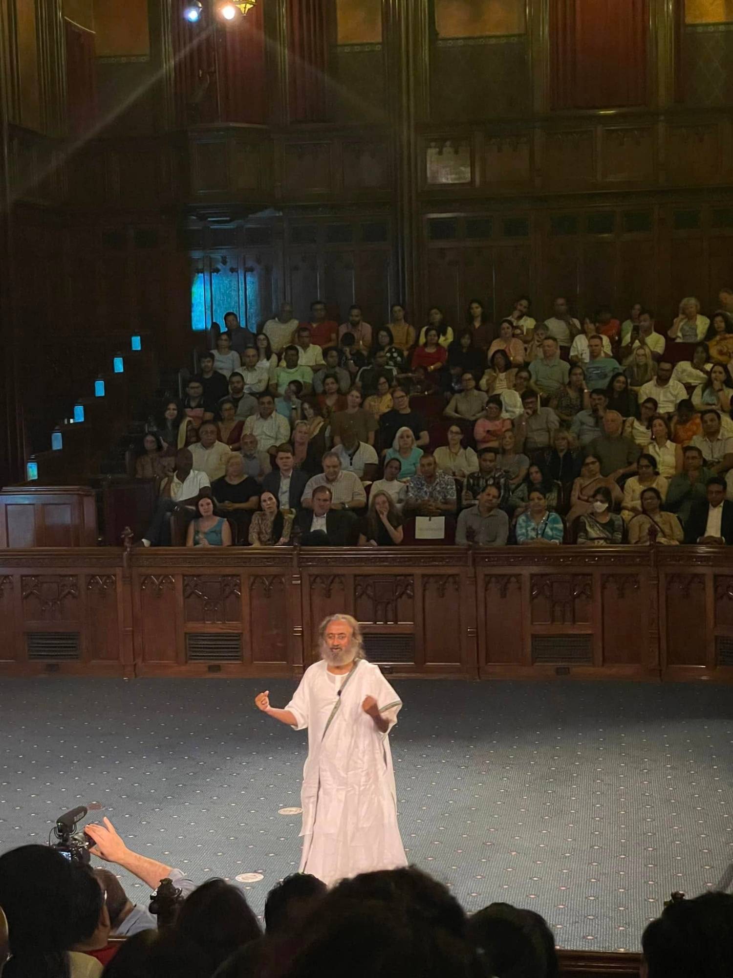 Gurudev Sri Sri Ravi Shankar speaking from the floor of a wood-paneled hall to a packed audience