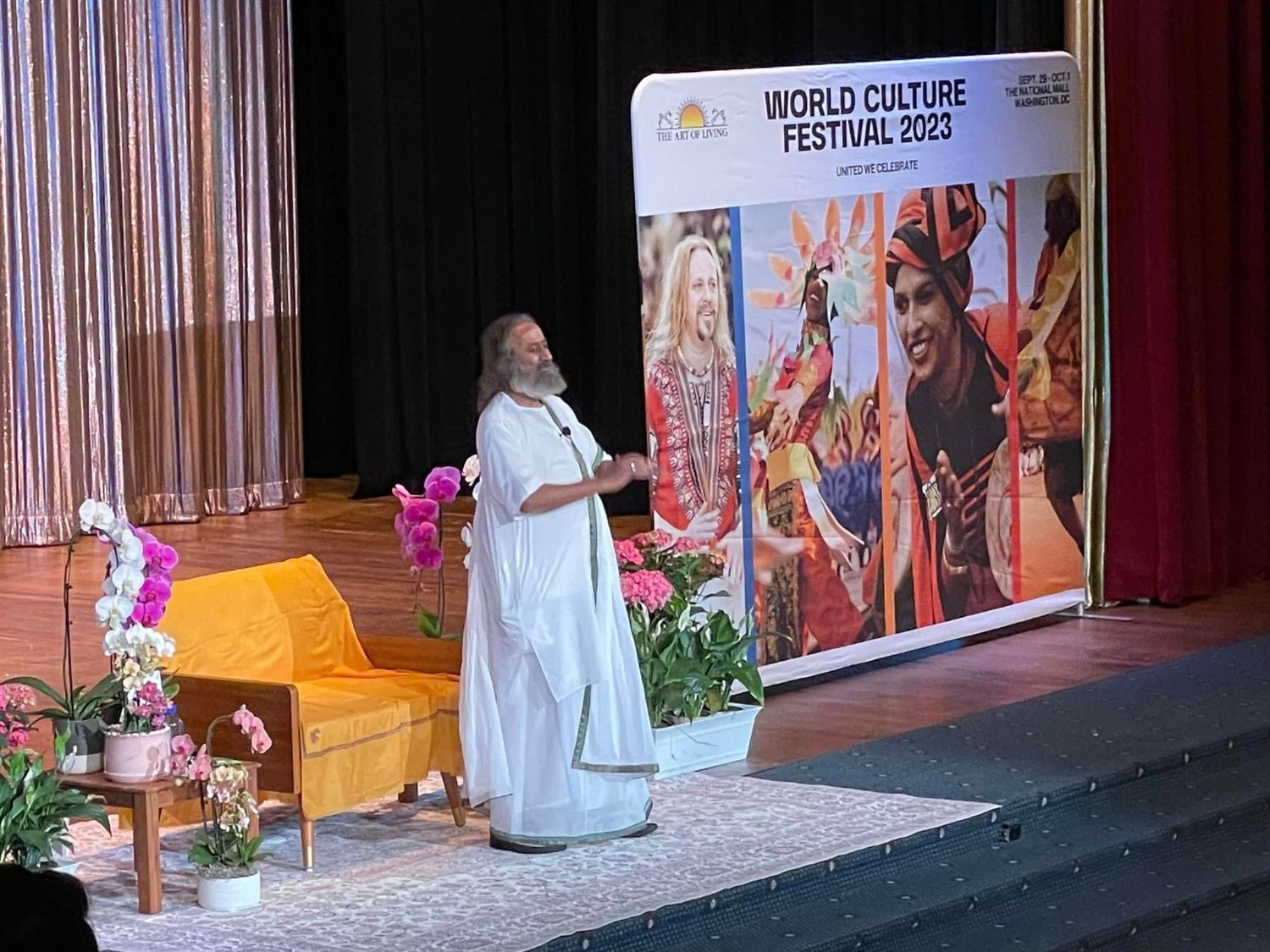Gurudev Sri Sri Ravi Shankar, dressed in white, on stage beside orchids, a saffron-draped chair, and a World Culture Festival 2023 banner